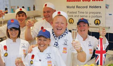 A smiling team of people in the fish and chip shop celebrate with glasses of champagne.
