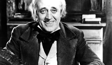 Image shows Scrooge smiling and writing with a quill.