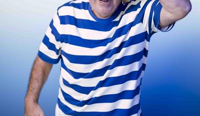 Image shows Danny Baker dressed as a sailor looking at the camera with his arm raised to shade his eyes as he looks.