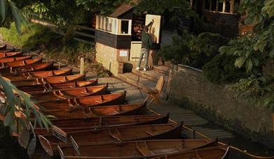 Boats lined up at the river bank. An attendant opens the door of a wooden hut.