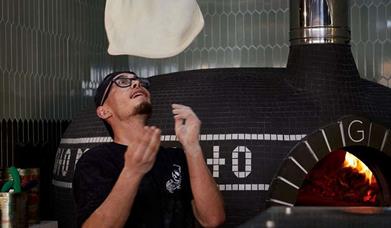 Moto pizza chef, throwing the pizza dough in the air with a pizza oven in the background