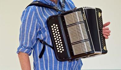 Image shows John Kirkpatrick smiling at the camera with an accordion