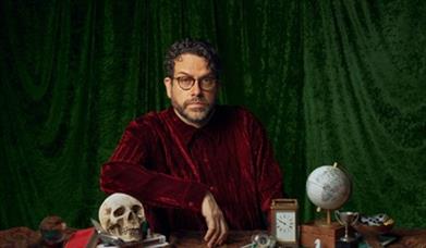 Image shows Michael Spicer looking serious with a skull next to him, a clock and a globe.